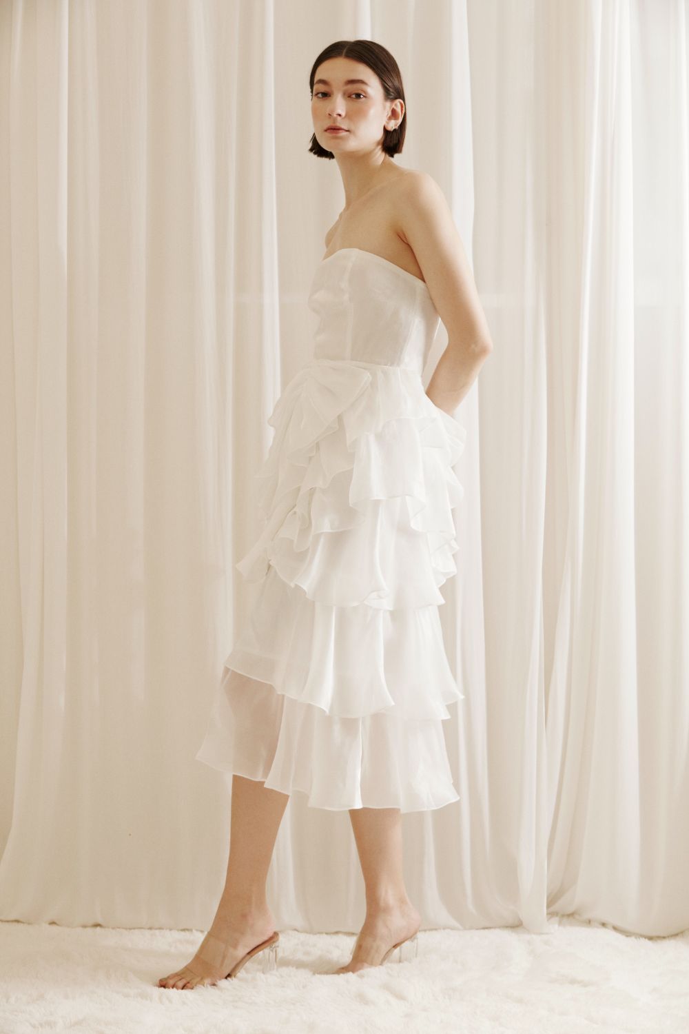 Side view of a woman wearing a white strapless dress with asymmetrical ruffled layers against a white curtain background