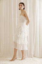 Side view of a woman wearing a white strapless dress with asymmetrical ruffled layers against a white curtain background