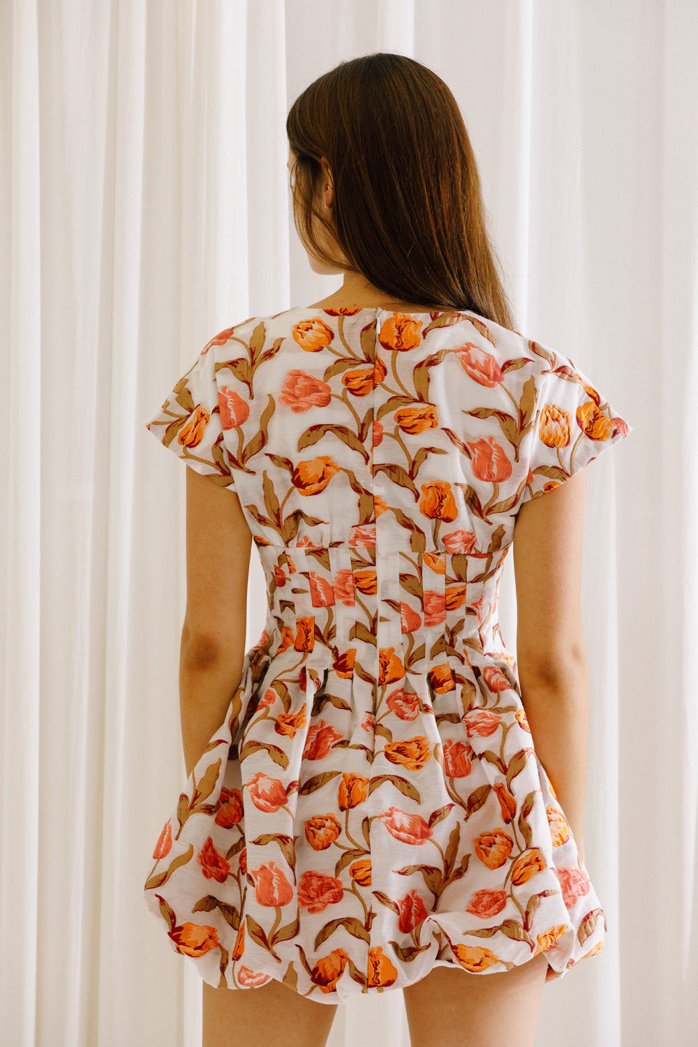 Back view of a woman wearing a coral and white floral dress against a white curtain background