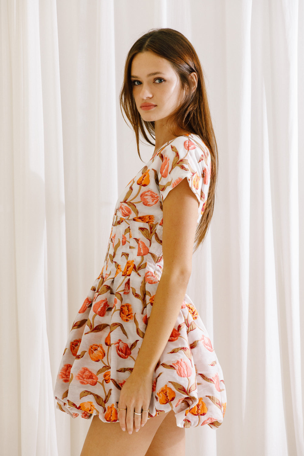 Side view of a woman wearing a coral and white floral dress against a white curtain background