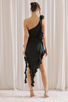 Back view of a woman wearing a black one-shoulder dress with ruffles against a white curtain background