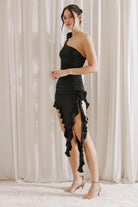 Side view of a woman wearing a black one-shoulder dress with ruffles against a white curtain background