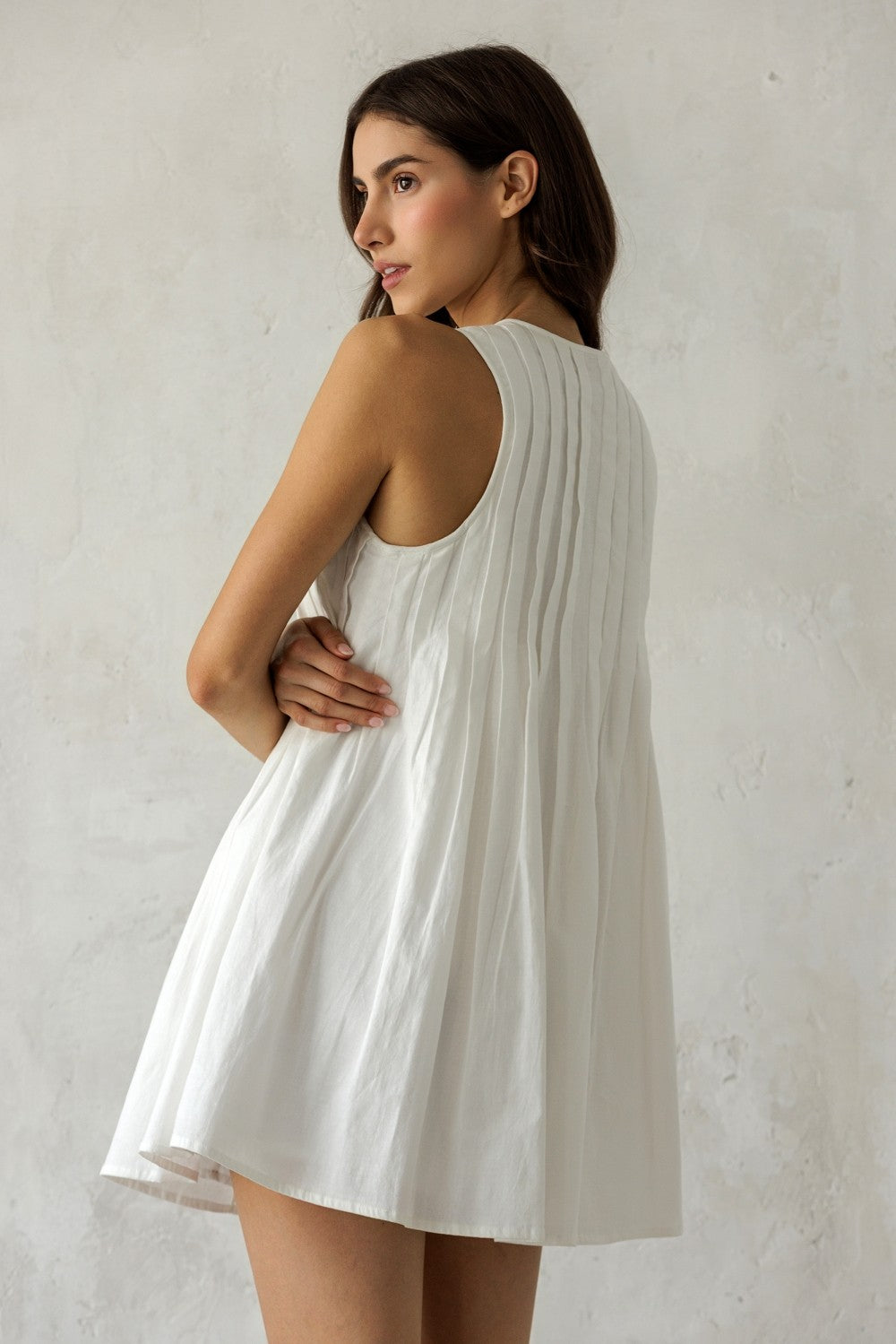 Back of a woman wearing a white sleeveless dress with a pleated design against a plain background