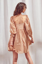 Back view of a woman wearing a brown dress with shimmering gold flaked embroidered dots and an invisible zipper against a white curtain background
