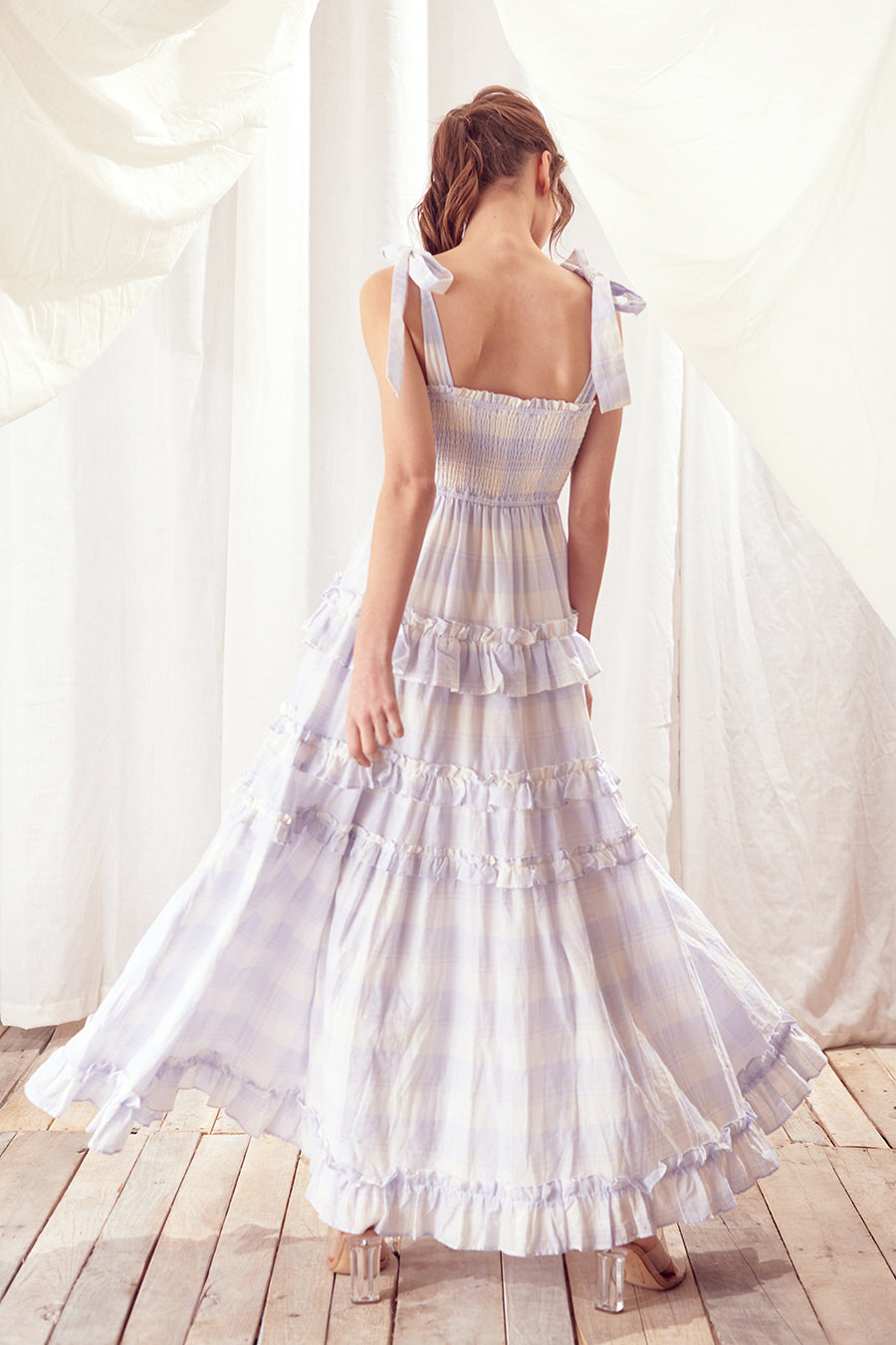 Back view of a woman wearing a white dress and blue plaid against a white curtain background