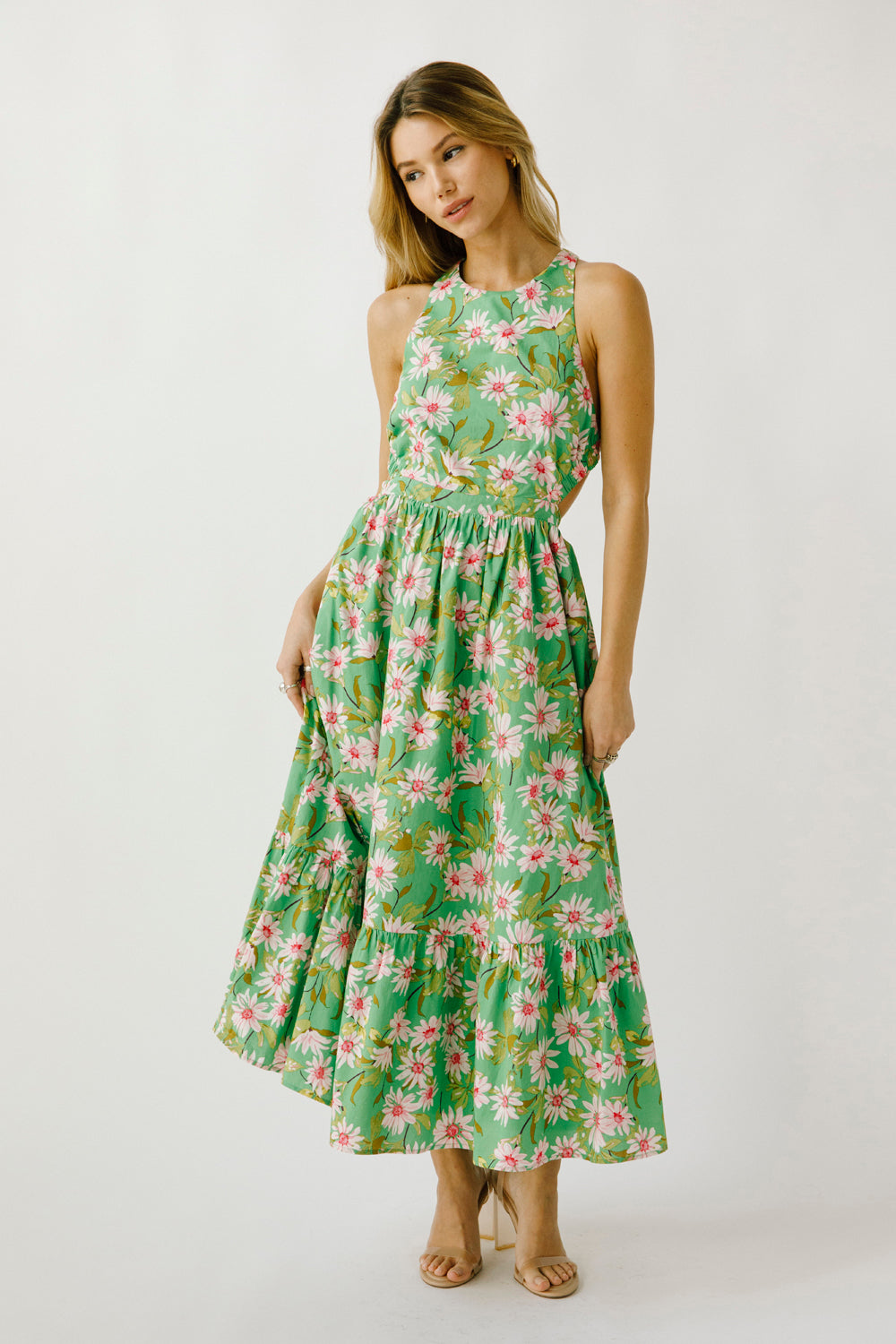 A model wearing a sleeveless, green maxi dress with a floral pattern, an elastic waistband, and a ruffle flowy hem.