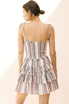 Back view of a woman wearing a gray and white tiered striped dress with self-tie thin straps on a plain background