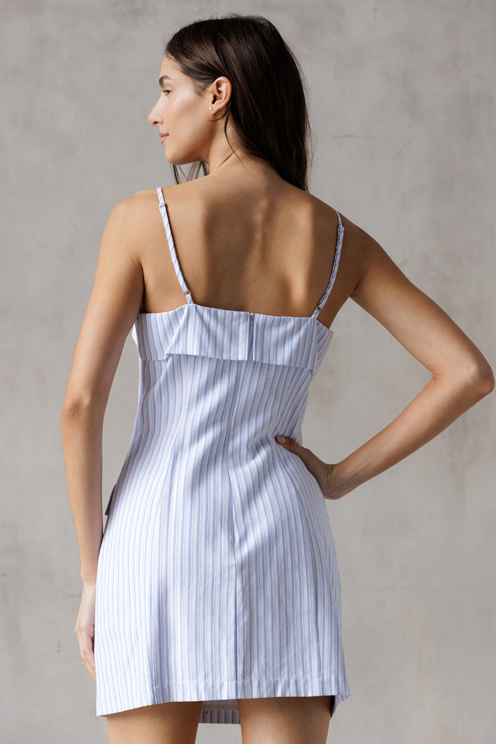 Back view of a woman wearing a blue and white striped dress with thin adjustable straps against a gray background