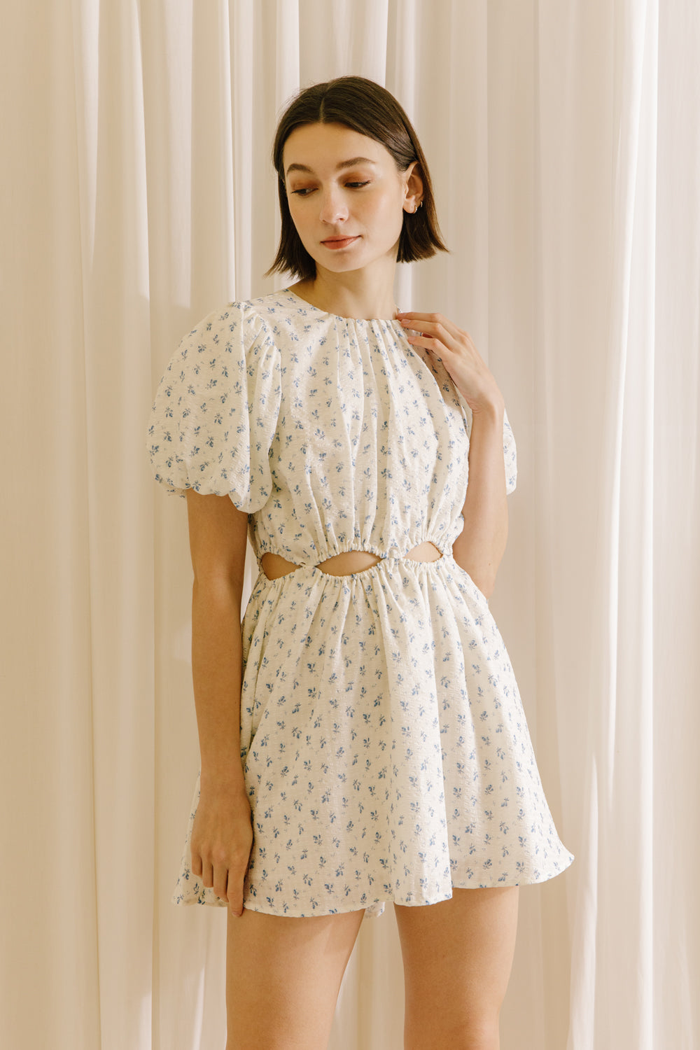 Woman wearing a cream dress with a floral pattern and cut-outs to the waist against a beige curtain background
