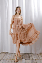 A model wears a dreamy warm-weather midi dress featuring a vintage floral print, square neckline, and flowy ruffled layers against a white curtain.