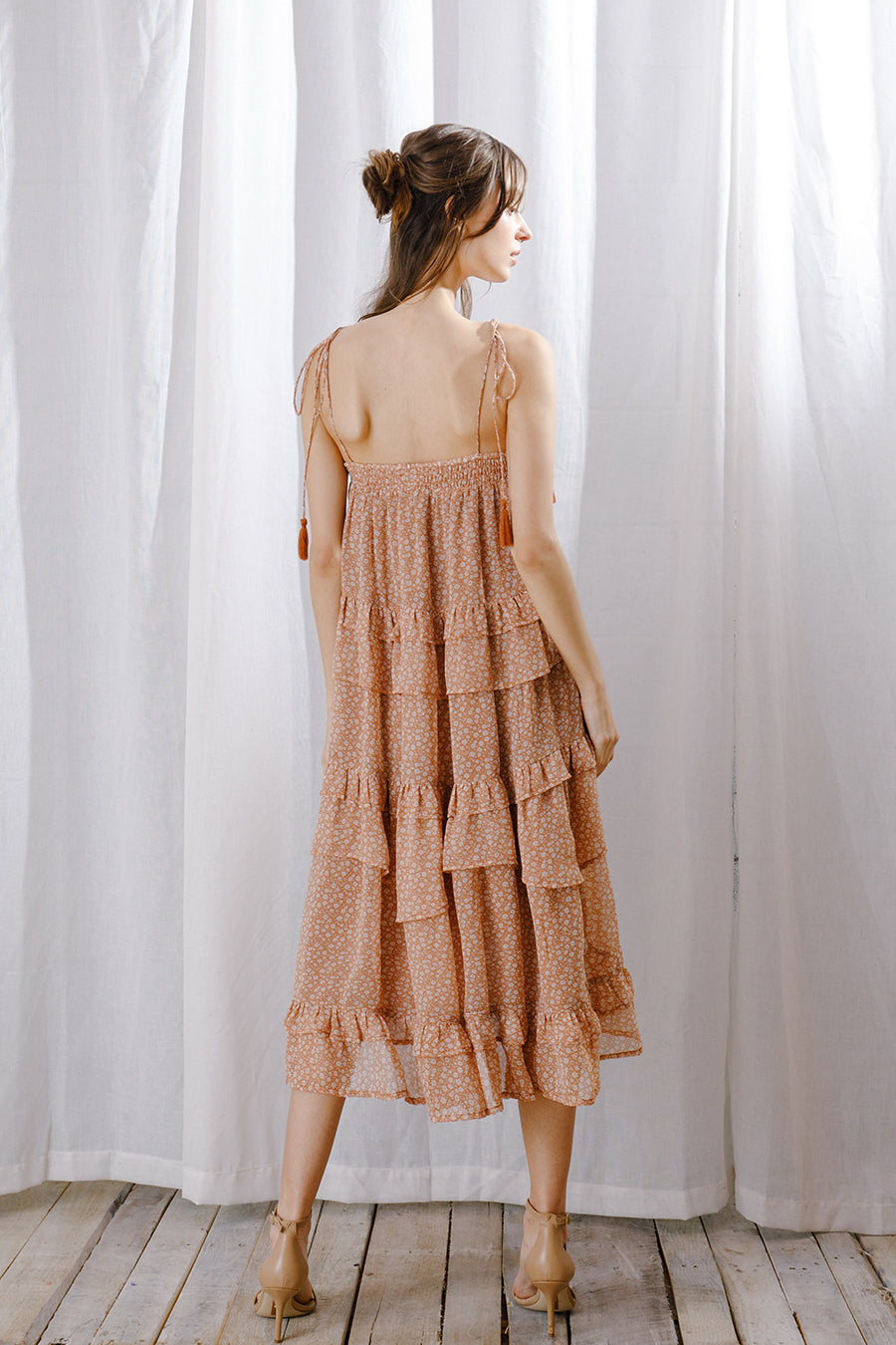 A woman viewed from behind wearing a rust floral midi dress with cascading ruffle layers, tassel tie straps, and a relaxed flowy fit.