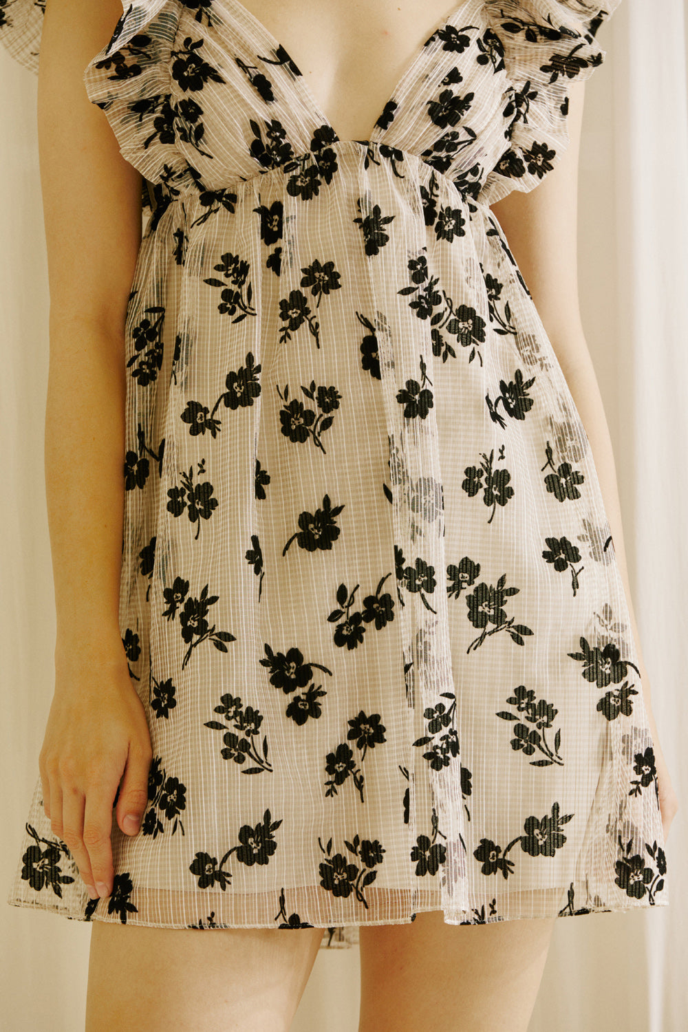 A person wearing a beige dress with a black floral sheer print, v-neckline and ruffled cap sleeves against a plain background