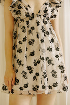 A person wearing a beige dress with a black floral sheer print, v-neckline and ruffled cap sleeves against a plain background