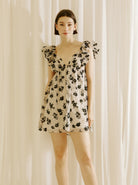Woman wearing a beige dress with black floral sheer print, v-neckline and ruffled cap sleeves against a plain background