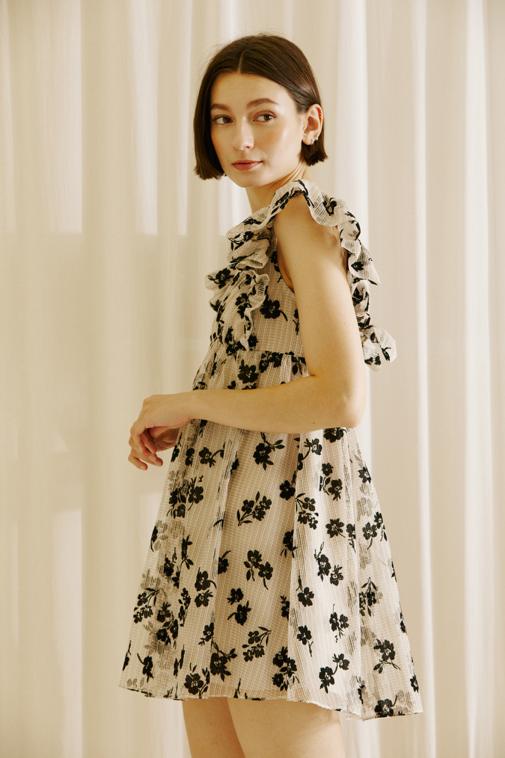 Side view of a woman wearing a beige dress with black floral sheer print, v-neckline and ruffled cap sleeves against a plain background