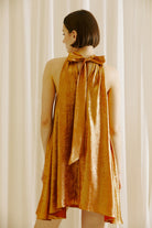 Back view of a woman wearing a bronze halter neck dress against a white curtain background