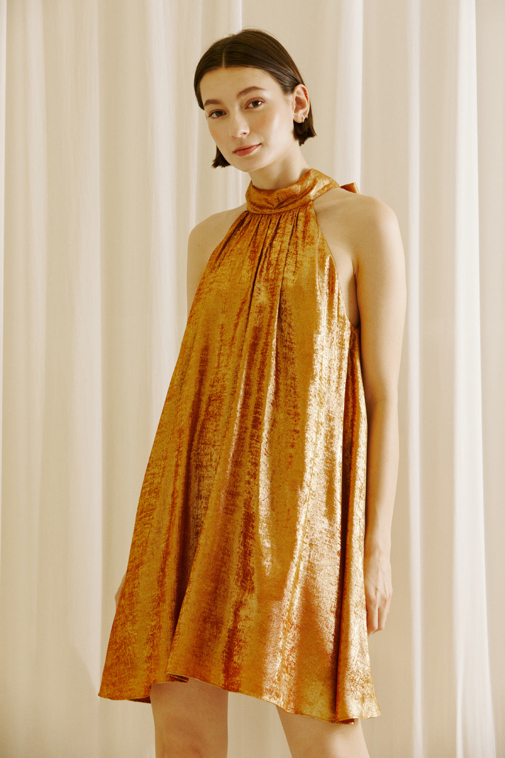 Woman wearing a bronze halter neck dress against a white curtain background
