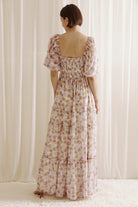 Back view of a woman wearing a purple floral dress with a slit standing against a white curtain.