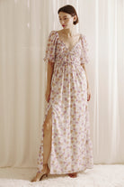 Woman wearing a purple floral dress with a slit standing against a white curtain.