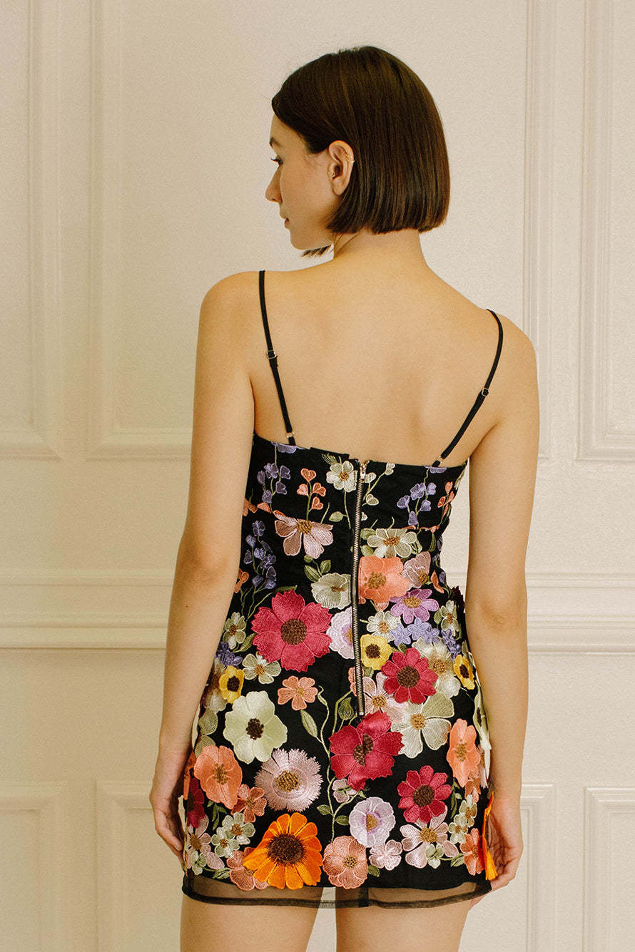 Back view of a woman wearing a black dress with colorful floral embroidery against a neutral wall.