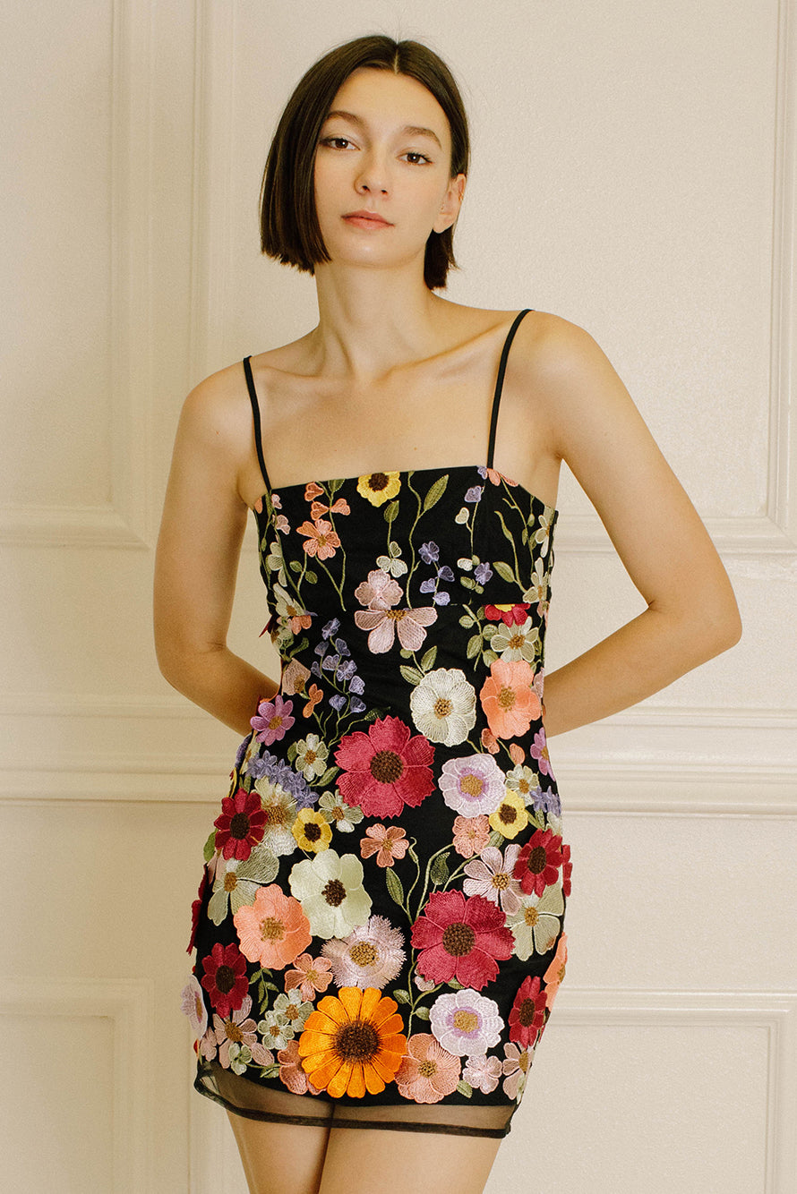 Woman wearing a black dress with colorful floral embroidery against a neutral wall.