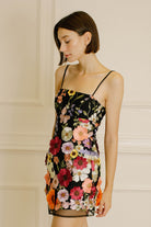 Side view of a woman wearing a black dress with colorful floral embroidery against a neutral wall.