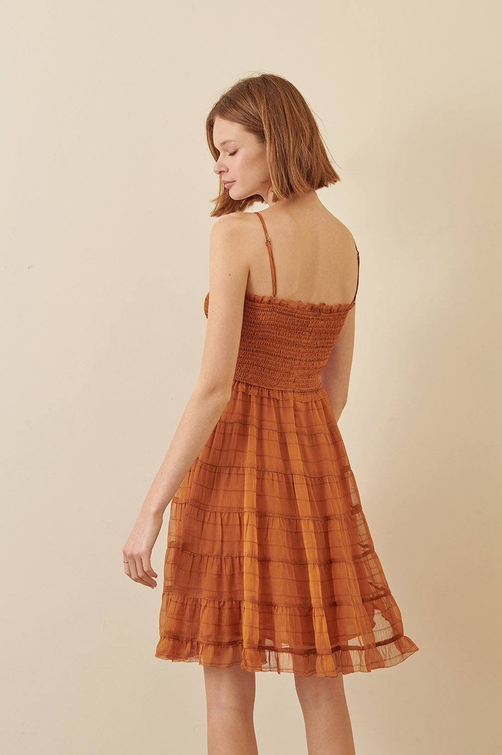 Back view of a woman wearing a rust, ruffled cami dress with spaghetti straps and a straight neckline.