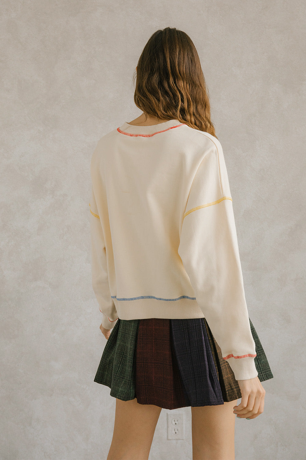 Back view of a woman wearing a cream pullover sweater with long sleeves and pleated plaid skirt. It has a crewneck and an oversized, loose fit. Sweet en Forme