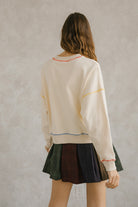 Back view of a woman wearing a cream pullover sweater with long sleeves and pleated plaid skirt. It has a crewneck and an oversized, loose fit. Sweet en Forme