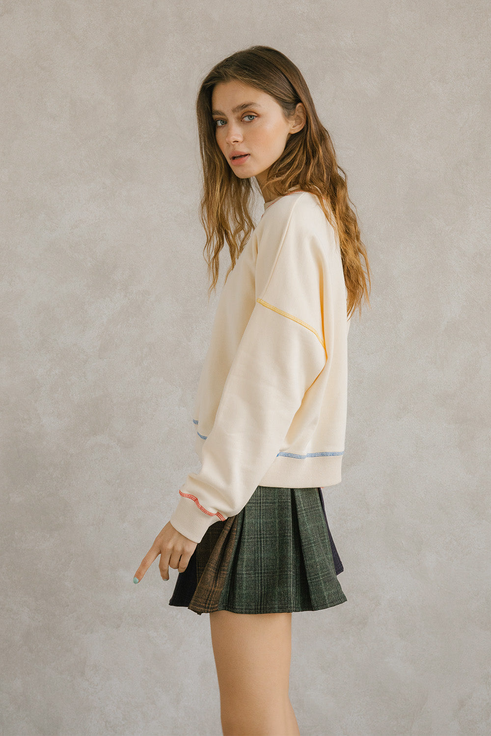 Side view of a woman wearing a cream pullover sweater with long sleeves and pleated plaid skirt. It has a crewneck and an oversized, loose fit. Sweet en Forme