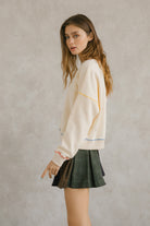 Side view of a woman wearing a cream pullover sweater with long sleeves and pleated plaid skirt. It has a crewneck and an oversized, loose fit. Sweet en Forme