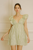 A woman wearing a short, sleeveless, green floral mini dress with a deep v-neckline and ruffled detailing.