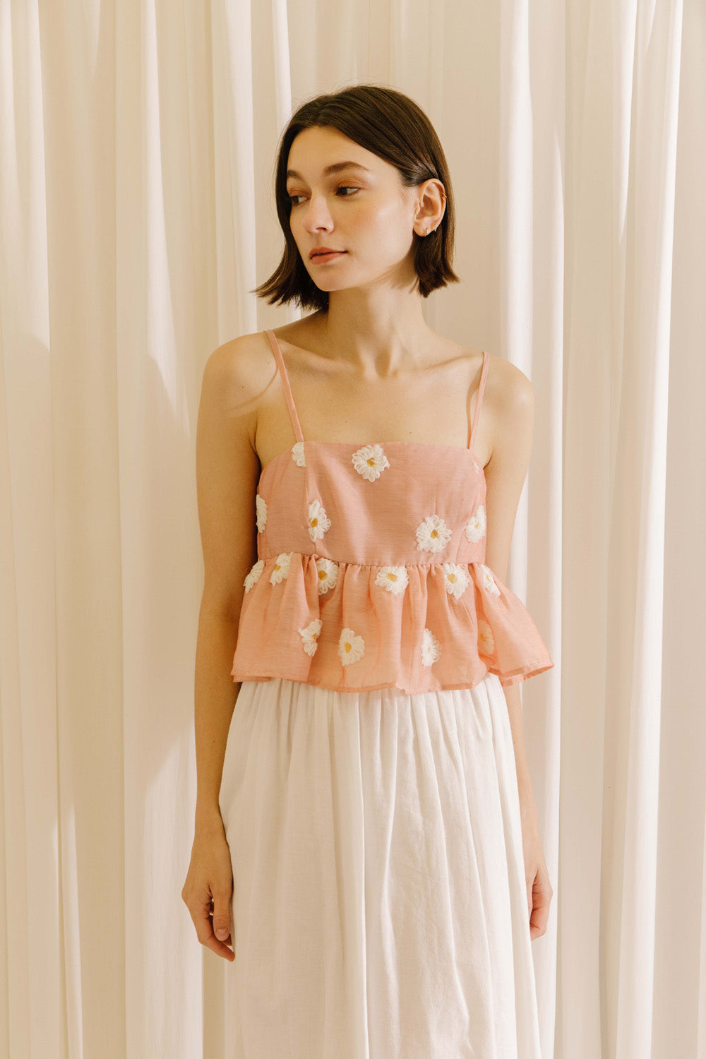 Woman wearing a pink floral cropped top and white pants against a beige curtain background