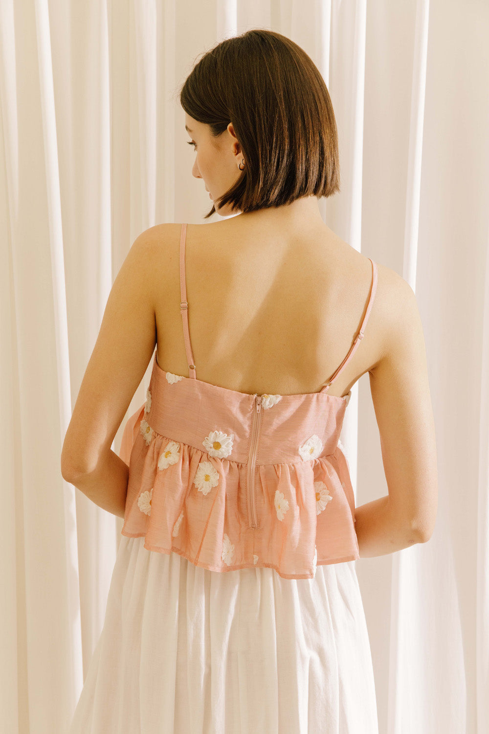 Back view of a woman wearing a pink floral top and white pants against a beige curtain background