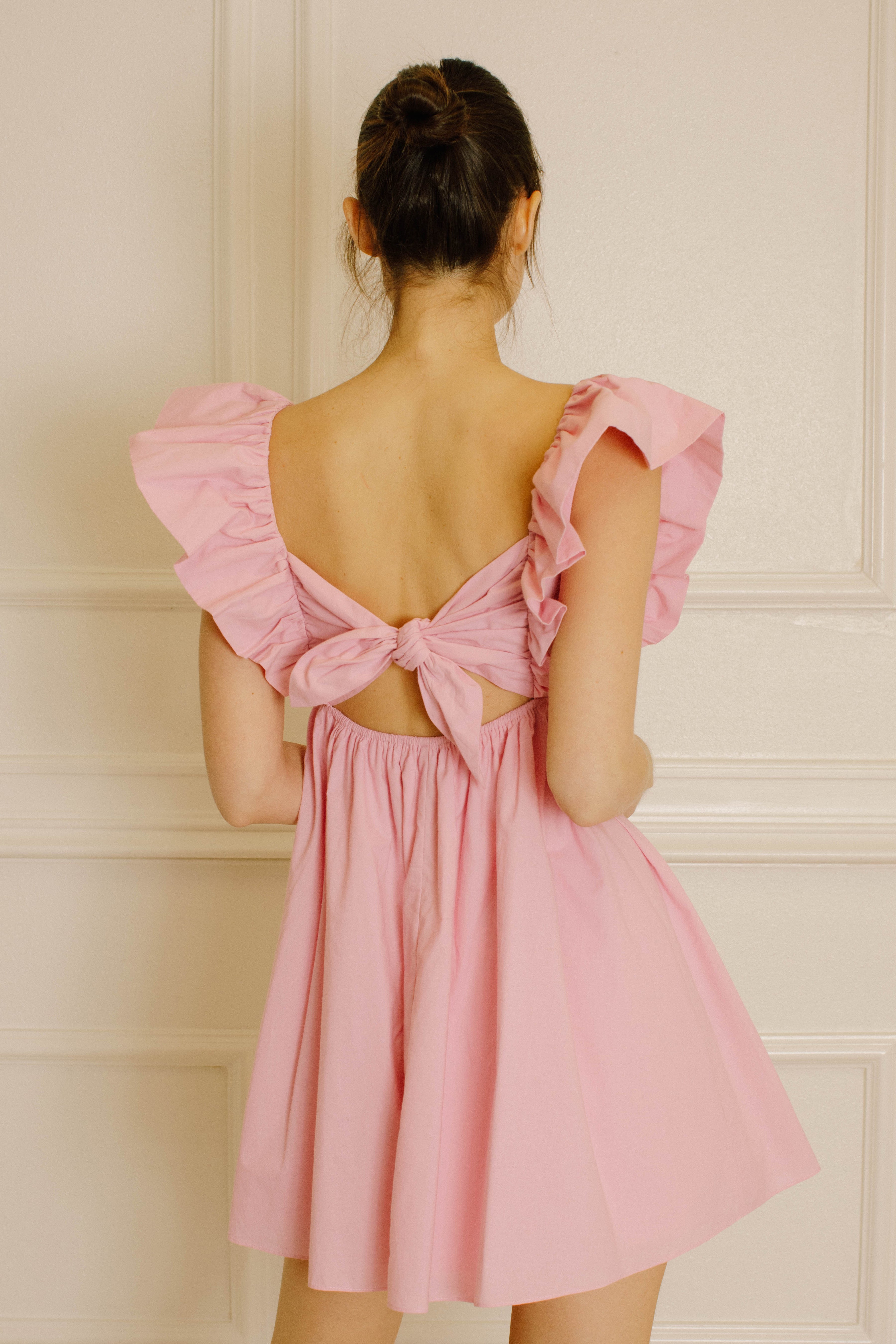 Back view of a woman wearing a pink dress with ruffled sleeves against a beige wall.