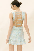 Back view of a woman standing wearing a floral white and blue mini dress with a crisscross back and ruffled detailing.