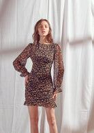 A woman modeling a black mini dress with a small orange and yellow floral pattern  standing against a white curtain