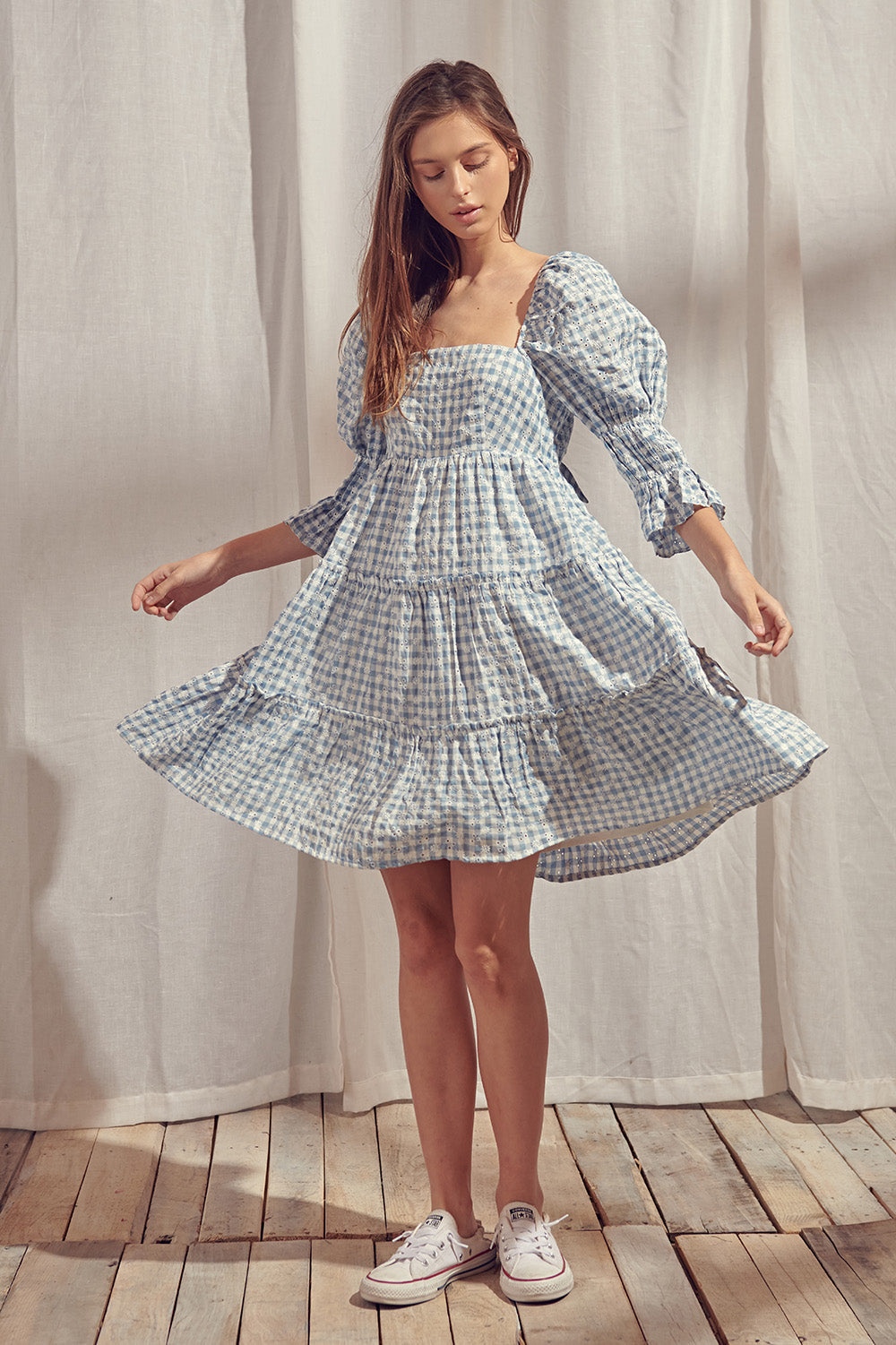 A woman wearing a blue and white gingham baby doll mini dress with a square neckline, 3/4 length puff sleeves, and an open back with a self-tie bow.