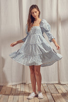 A woman wearing a blue and white gingham baby doll mini dress with a square neckline, 3/4 length puff sleeves, and an open back with a self-tie bow.