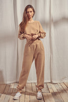 Woman wearing a beige sweater and pants standing against a white curtain.