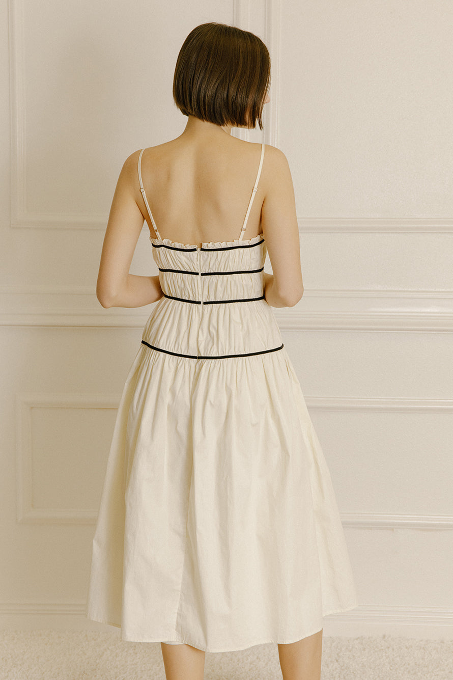 Back view of a woman wearing a cream midi dress with black trim against a neutral wall.
