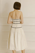 Back view of a woman wearing a cream midi dress with black trim against a neutral wall.