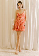 A woman standing in a white background wearing a vibrant coral 3D rose A-line mini dress with spaghetti straps and a cinched waist.