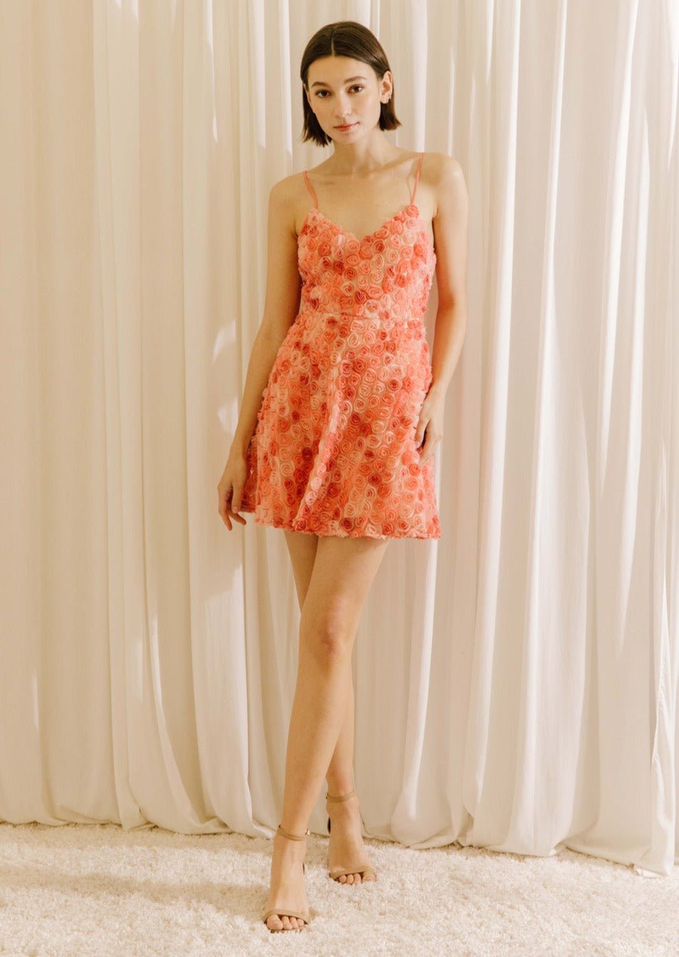 A woman standing in a white background wearing a vibrant coral 3D rose A-line mini dress with spaghetti straps and a cinched waist.