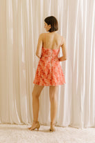 Back view of a woman standing in a white background wearing a vibrant coral 3D rose A-line mini dress with spaghetti straps and a cinched waist.