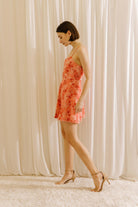 Side view of a woman standing in a white background wearing a vibrant coral 3D rose A-line mini dress with spaghetti straps and a cinched waist.