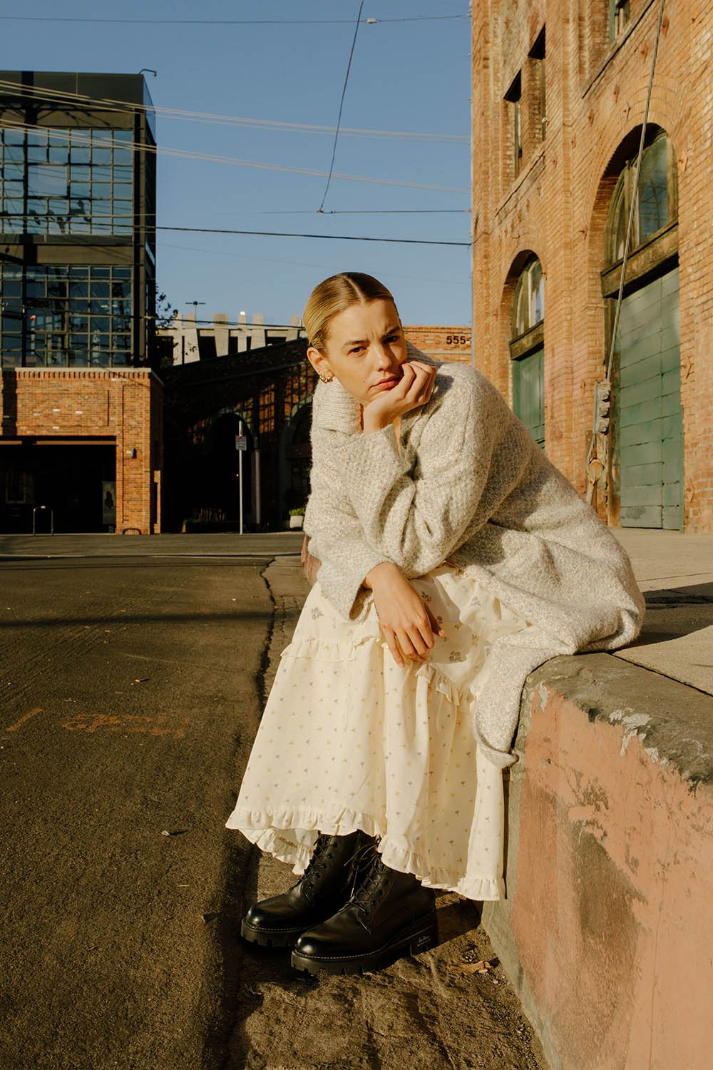 Woman in a long grey cardigan sitting on the sidewalk against a brick wall.