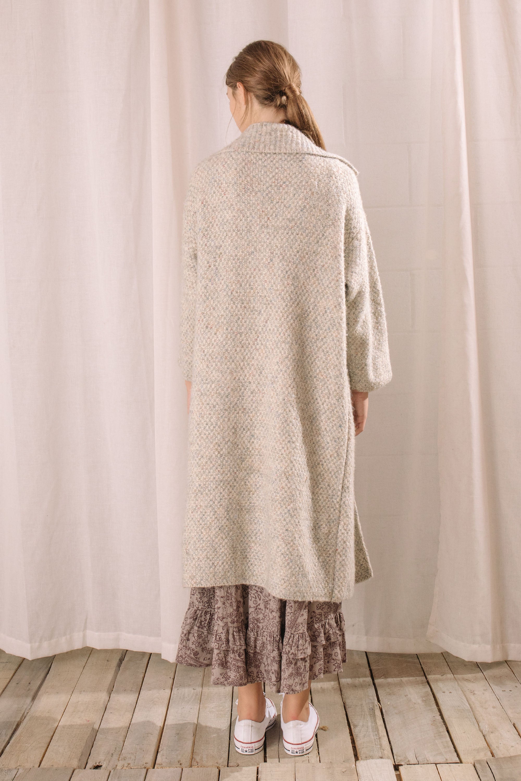 Back view of a woman in a long grey cardigan standing against a brick wall