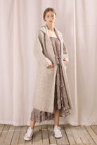 Front view of a woman in a long grey cardigan standing against a brick wall