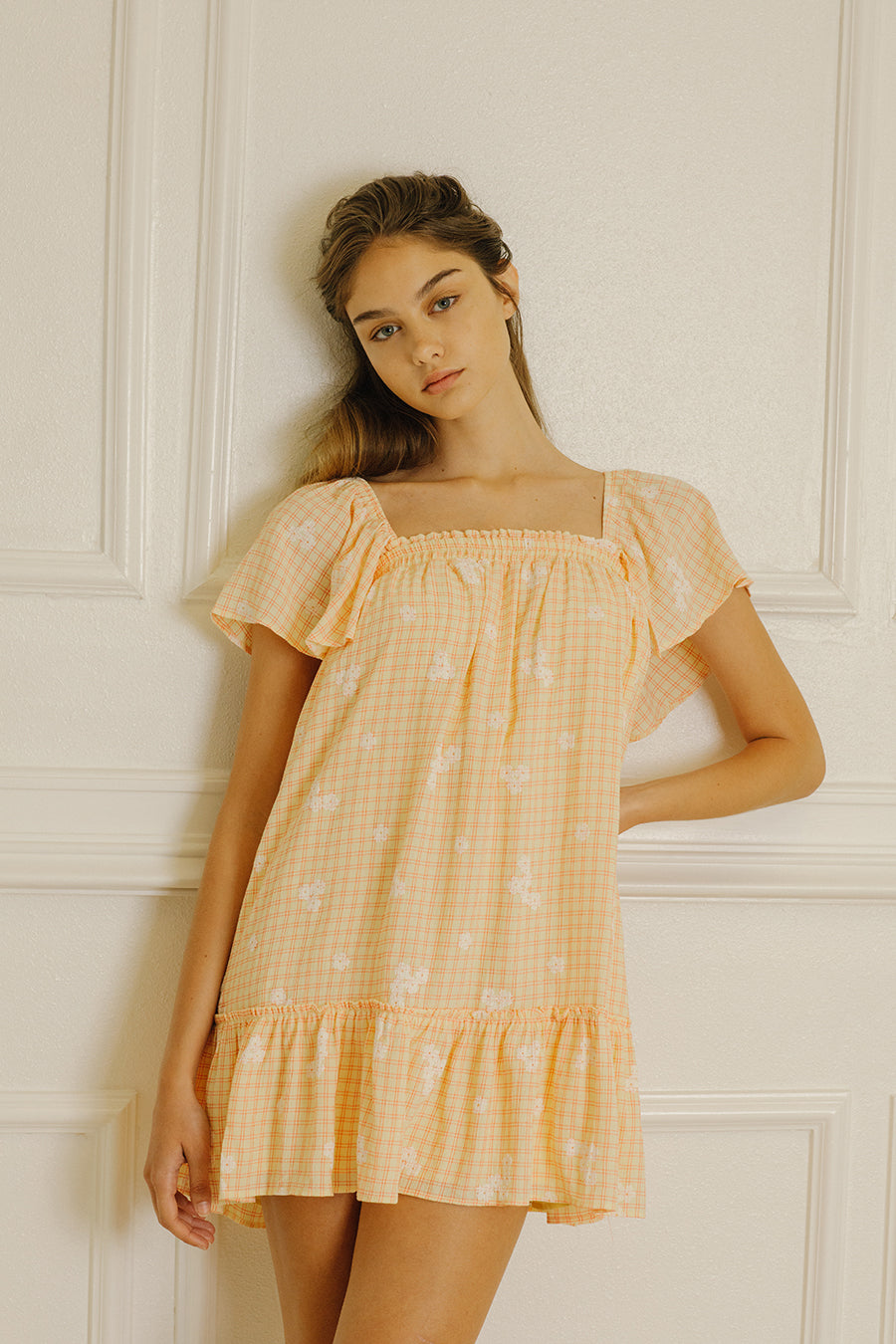 Woman in an yellow dress with a gingham and white daisy print against a neutral wall